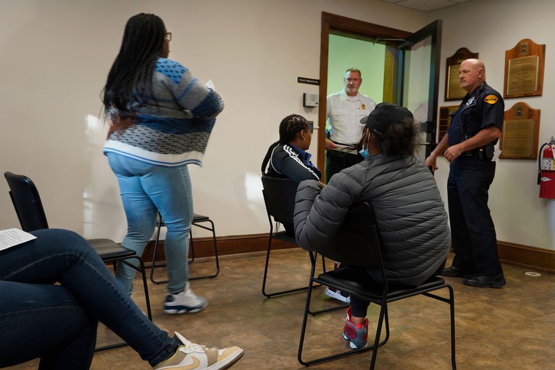 A Black woman walks out of a waiting room past three other Black women. Two White men in uniform stand at the front of the room by an open door.