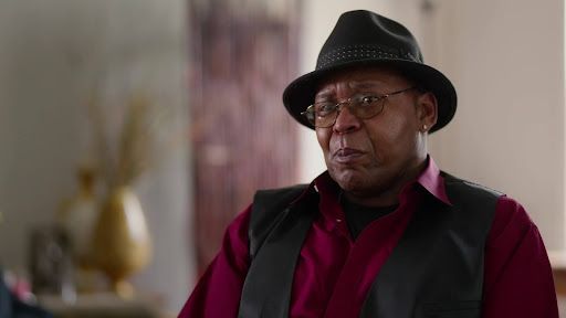 A Black man with glasses and a black fedora, black vest and red button-down shirt.