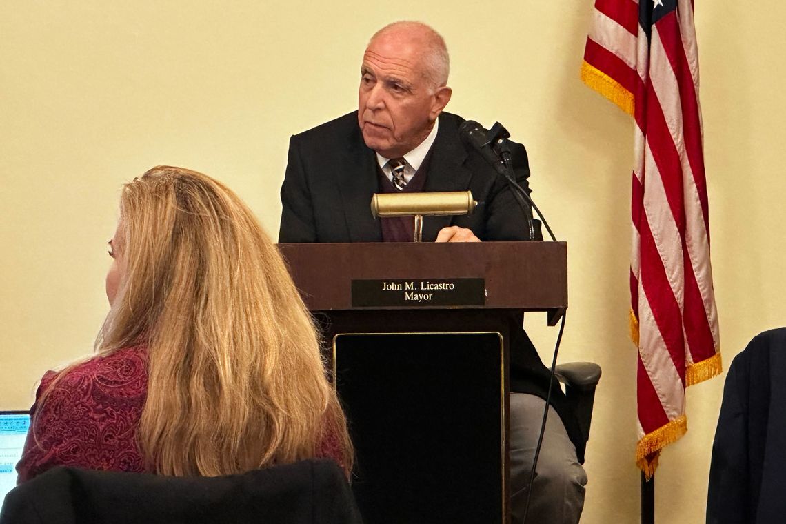 Mayor John Licastro, a White man with short white hair, sits behind a podium. A woman sits in front of him. 