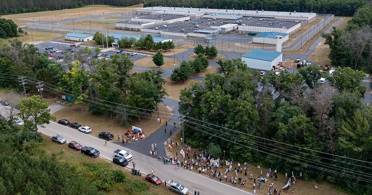 An aerial photo shows a prison facility composed of over half a dozen buildings, with trees surrounding much of the property. In the foreground, there are activists gathered with signs and banners near the facility.  