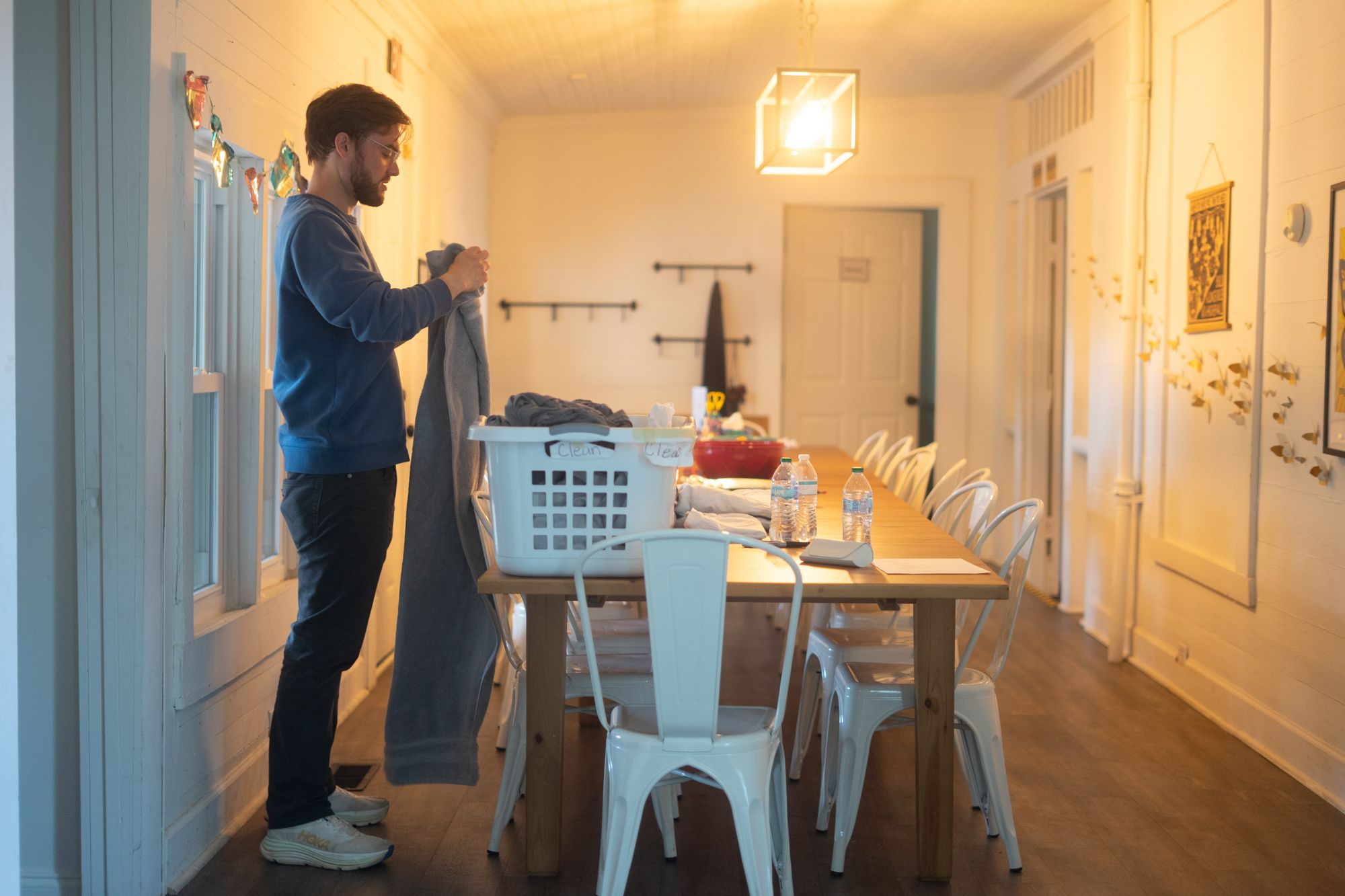 A man with light-toned skin, wearing a blue sweatshirt, holds a towel up in front of a basket of laundry that's sitting on a long table.