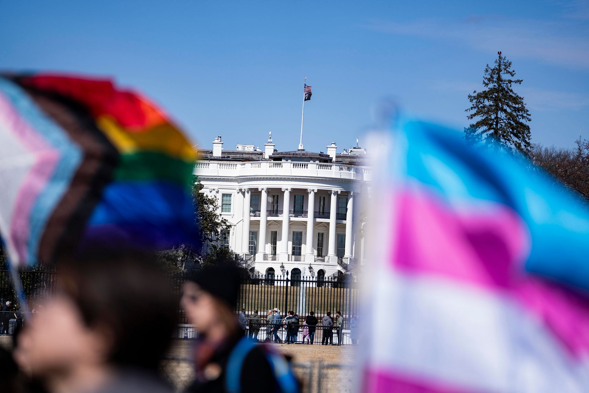 The White House, a white, multi-story building with columns and a flag on top, is visible between an out-of-focus rainbow-colored flag held up by protesters and an out-of-focus blue, pink and white flag.  