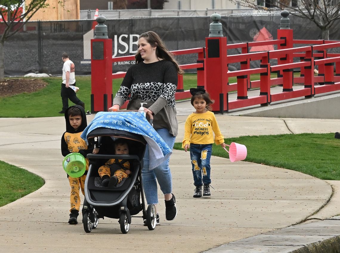 A light skin-toned woman pushes a stroller while walking through a park with her three children.