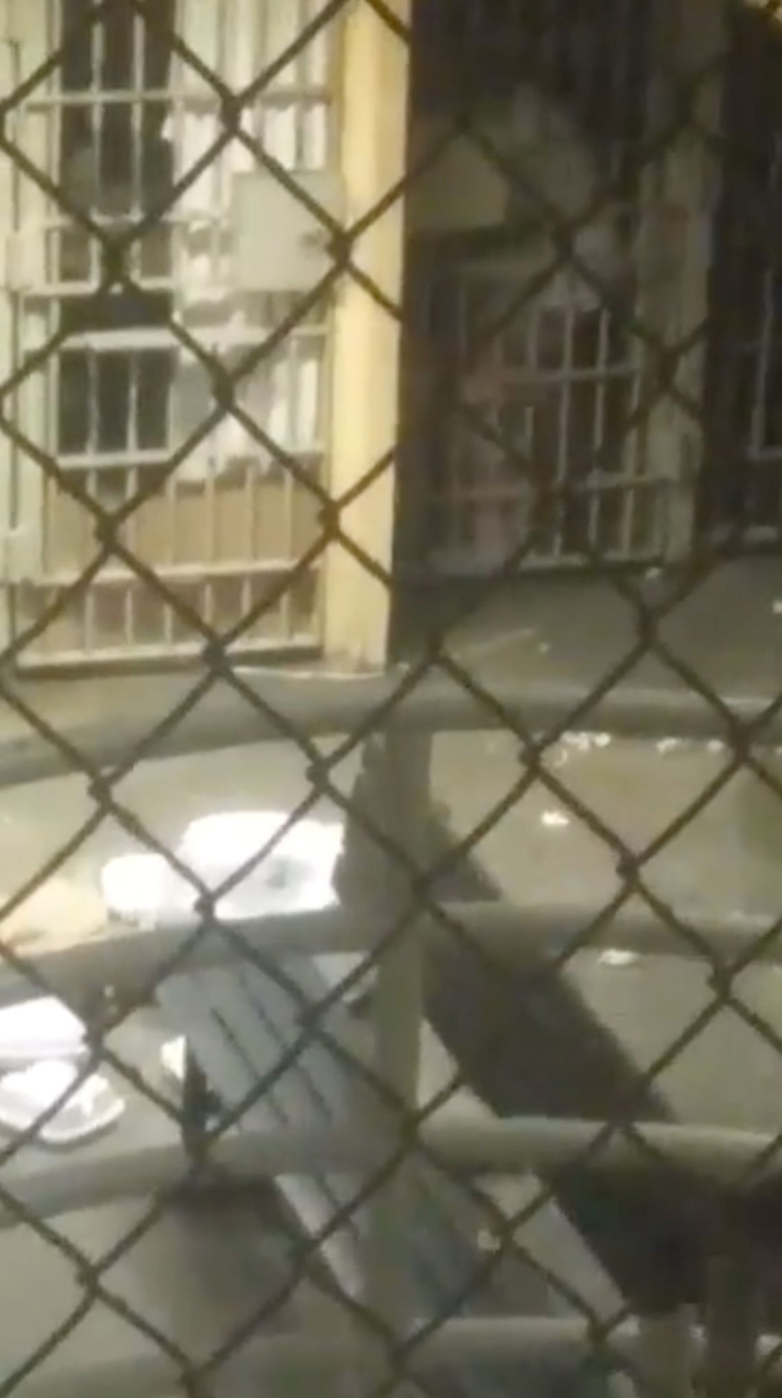 A blurry still image from a cell phone video filmed through a chainlink fence shows a person in a prison cell in darkness, while the rest of the unit is lit. 