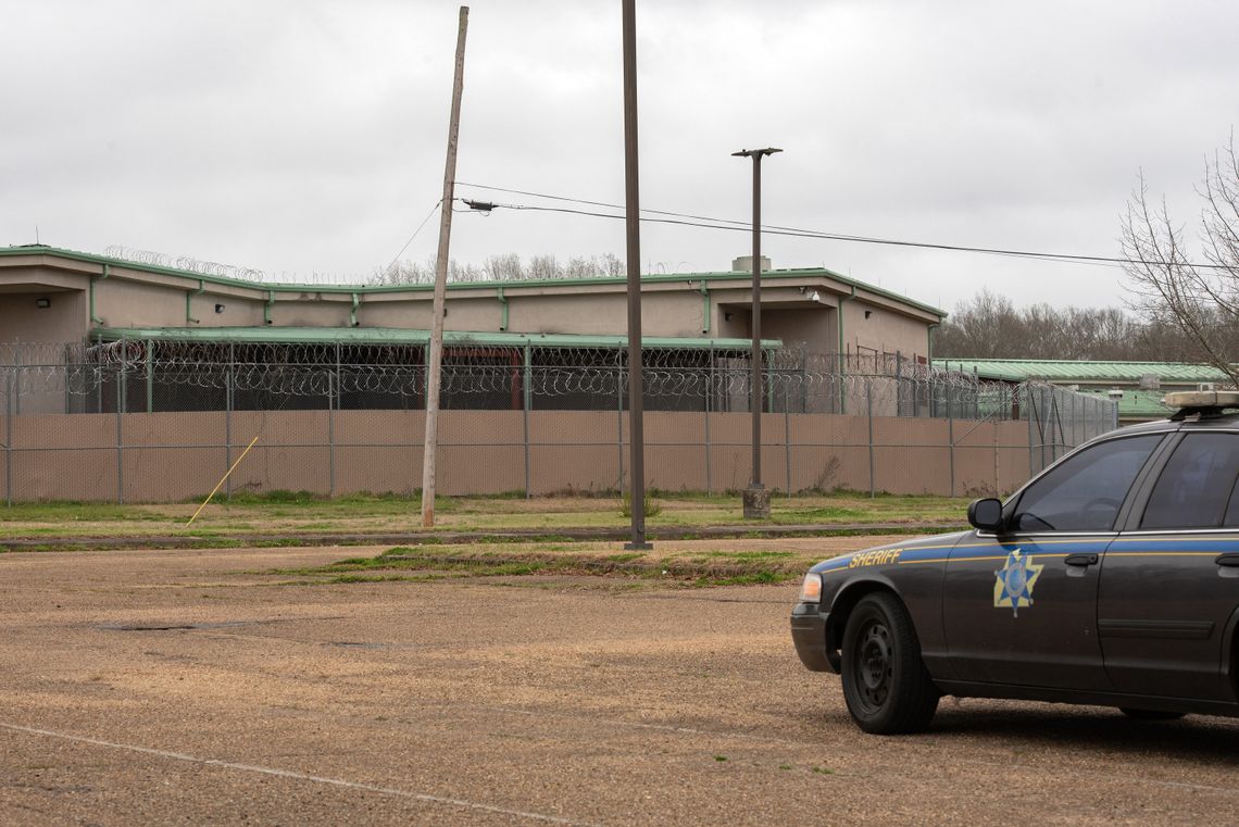 A car with the word “Sheriff” on it is parked on the right side of the photo, in front of a facility surrounded by a barbed wire fence. 