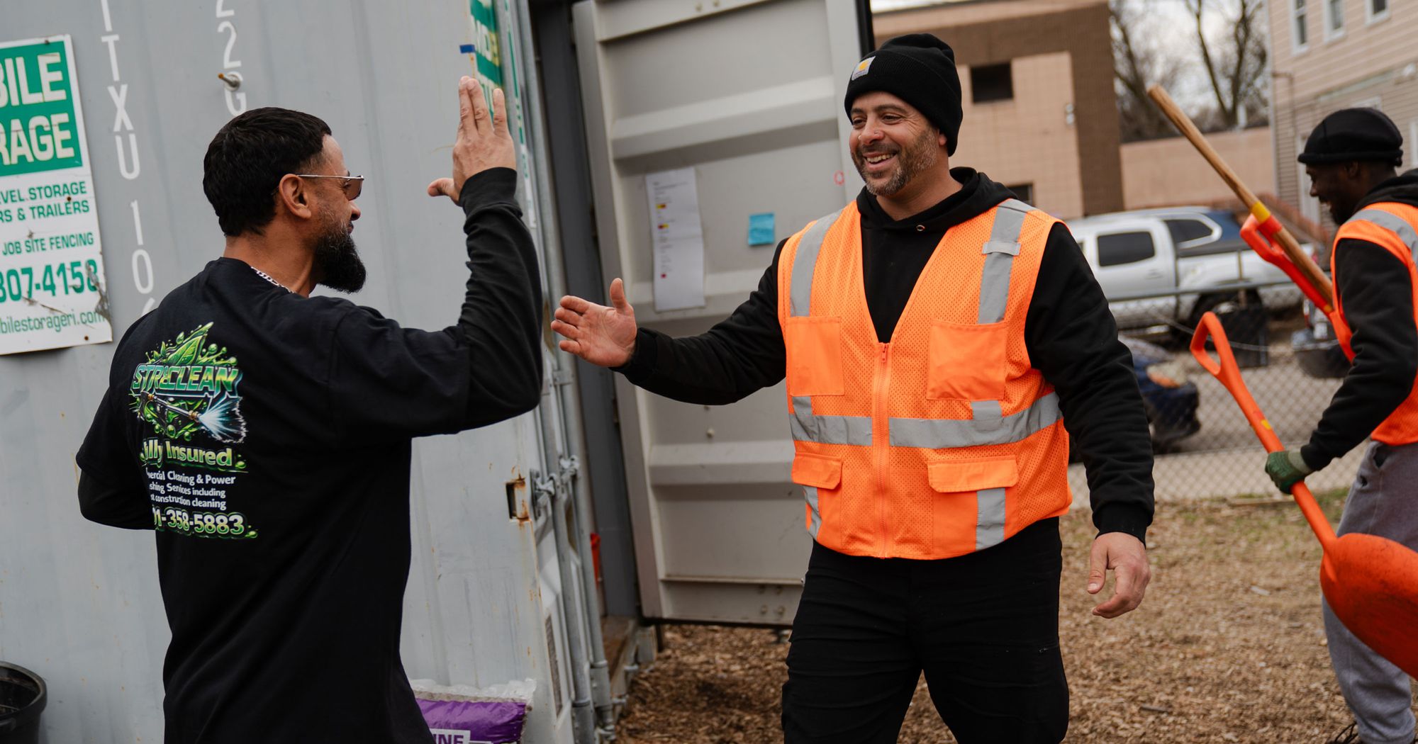 A photo of a man in an orange safety vest greeting another man in a black shirt. Both men have a medium skin tone and beards. 