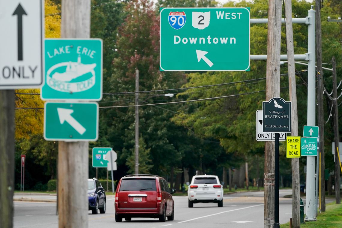 Road signs are visible in the foreground as a red van and white van enter the westbound ramp of I-90.