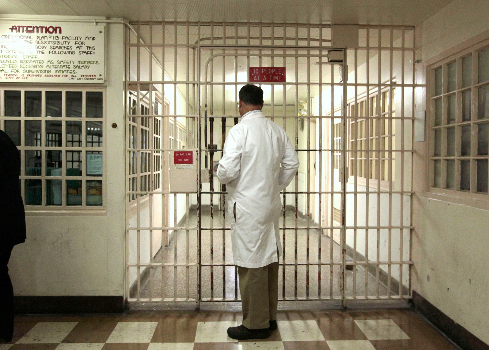 A male doctor, wearing a white lab coat and tan slacks, stands in front of a barred hallway.