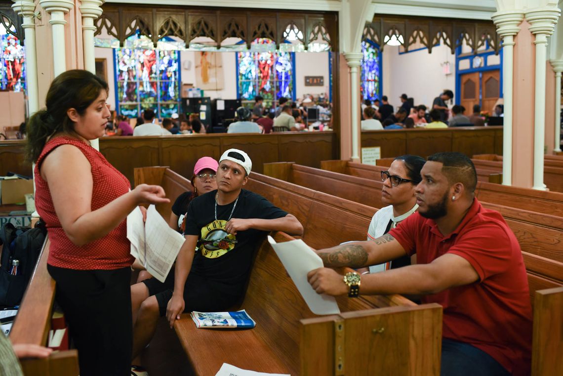 A group of medium-skinned people representing various genders sit in church pews, arms draped over the backs of the pews, while a medium-skinned woman with dark hair pulled back in a ponytail stands in front of them holding paperwork. A long row of stained-glass windows and a crowd of other people are in the background.