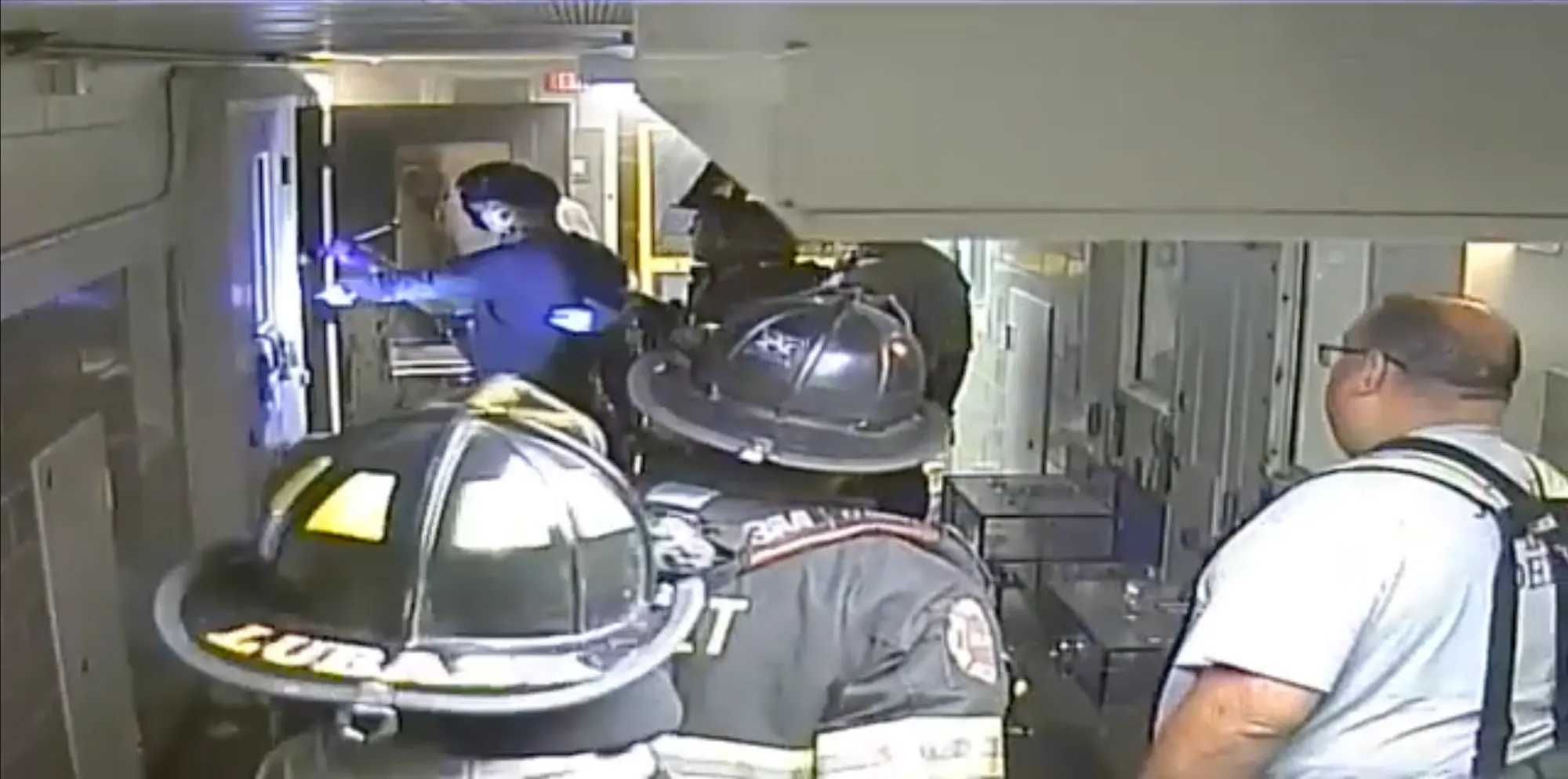 A video still shows firefighters examining a person lying down on the floor of a prison hallway. A cell door with the label 'A9' is open on the right. Several correctional officers stand nearby.