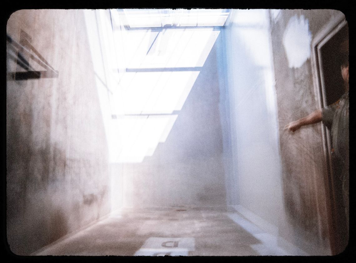 A photo shows an exercise space at the Solitary Housing Unit (SHU) at Pelican Bay State Prison. A man on the right of the photo is pointing his finger at the small space, which is confined by concrete walls. The photo has a double-vision effect achieved with a special lens. 