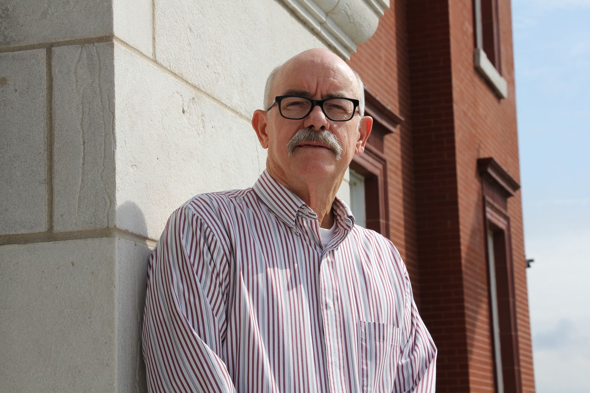 A photo shows a bald White man, wearing glasses and a striped button-down shirt, leaning against an exterior wall of a courthouse to pose for a portrait. 