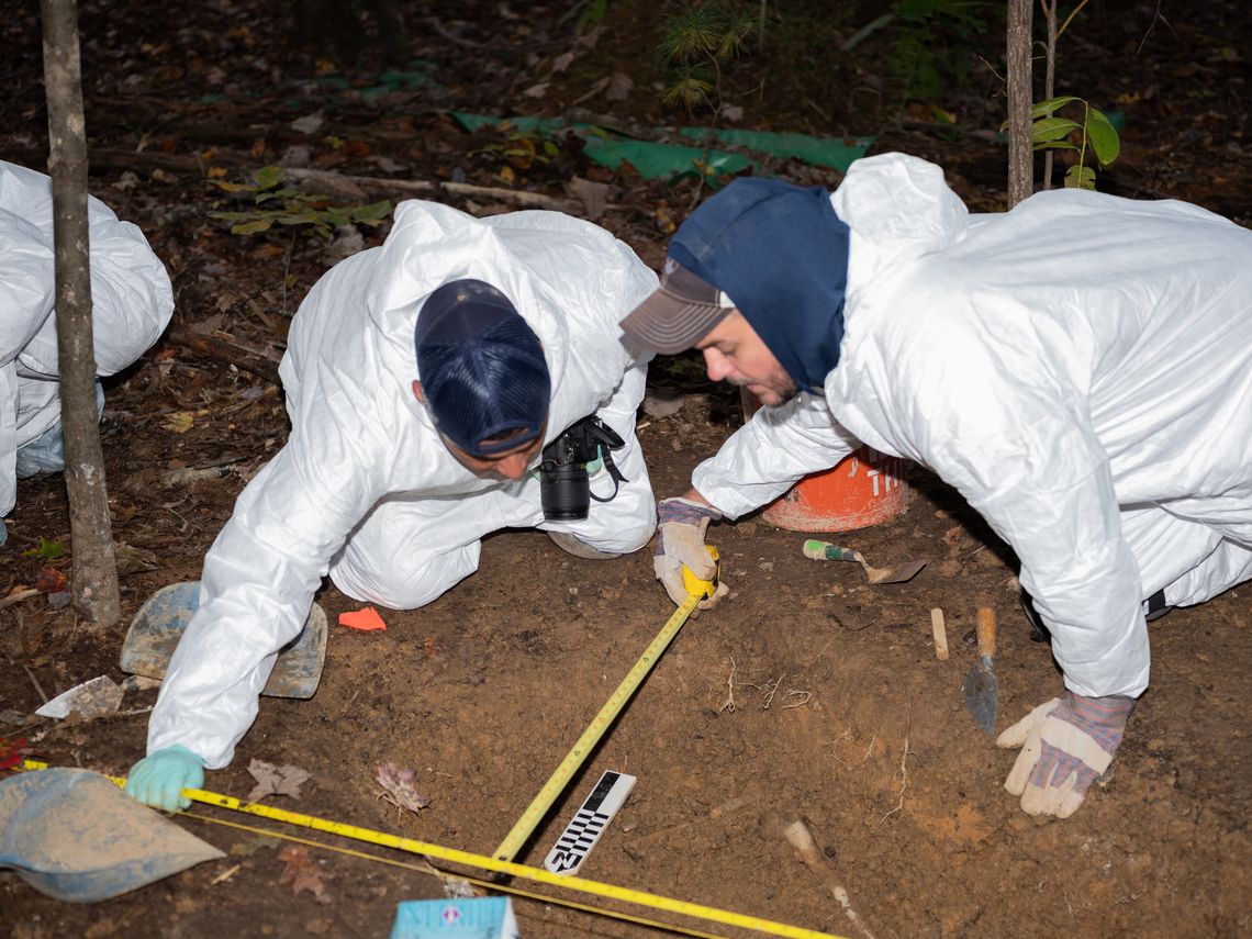 Two students dressed in hazmat suits use measuring tape to mark the location of human remains. 