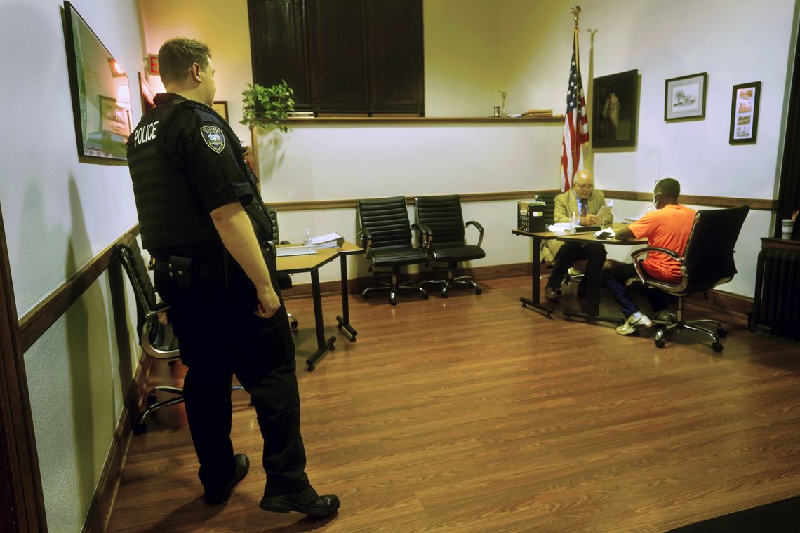 Prosecutor Tom Hanculak, a White man with short white hair, talks to a Black man in an orange T-shirt in a room as a White police officer in a uniform looks on. 