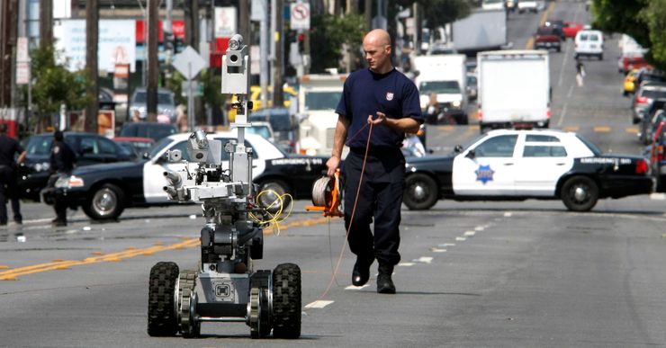 A robot operates in the middle of a street. A police officer walks behind it, while holding an extension cord reel connected to the robot. In the background, two police cars block the street from ongoing traffic. 