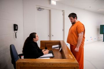 A White woman sitting behind a desk speaks to a White man standing up in a jail uniform. 