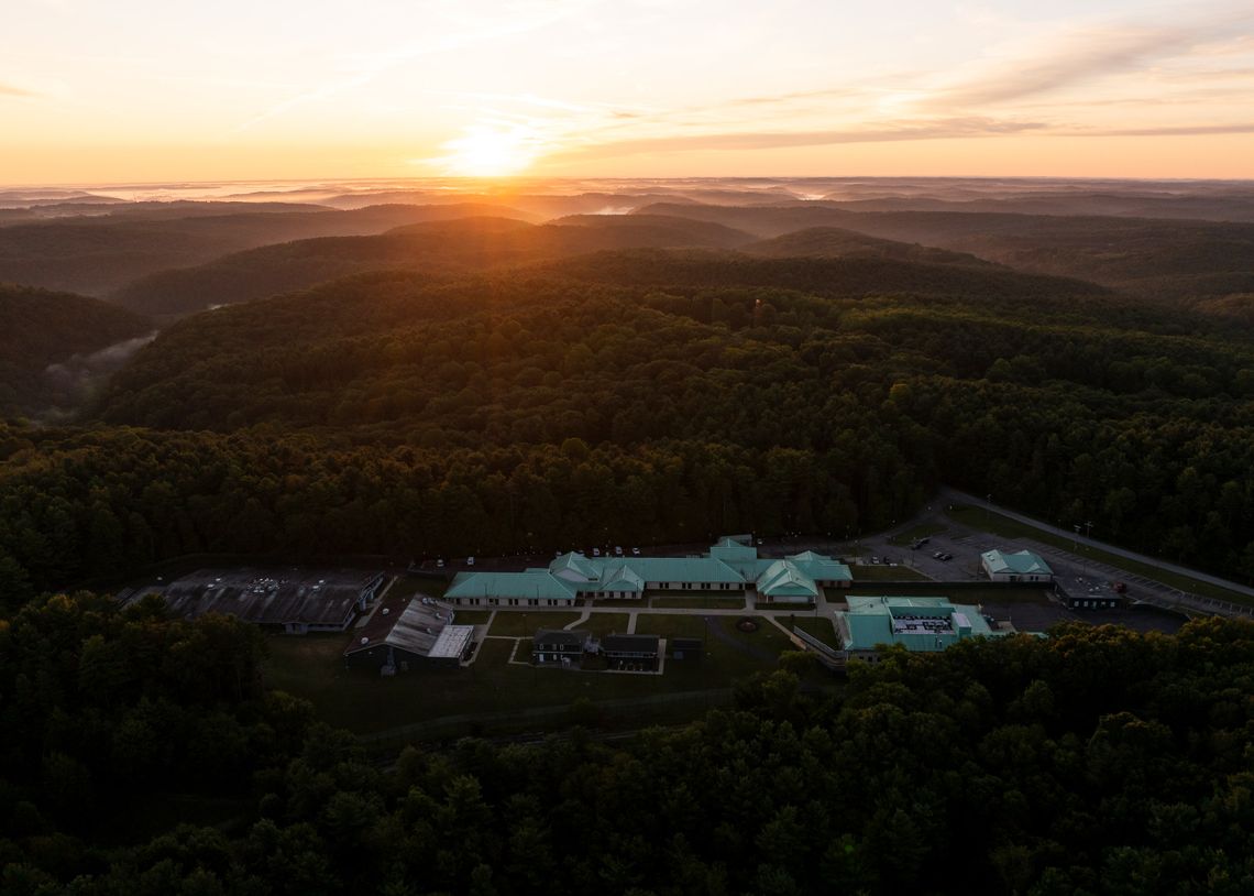 An aerial photo shows the campus of a residential treatment facility with a light green roof during sunset. The facility is surrounded by forest and hills.