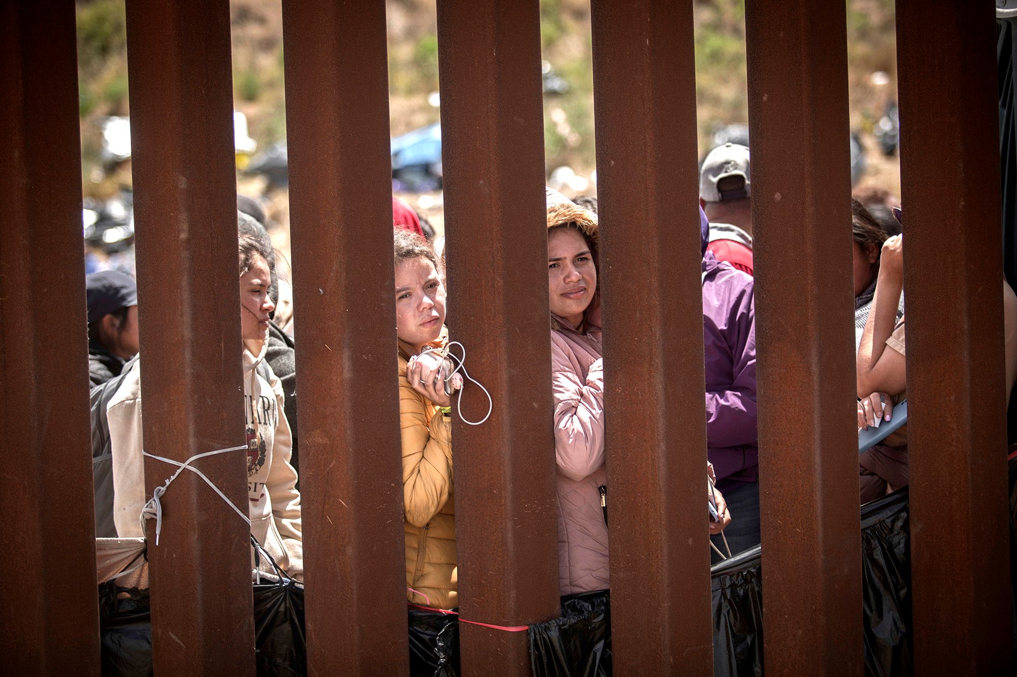 Two women with medium skin tone, holding onto their belongings, stand in between brown pillars of a wall while waiting in a line. Several people stand in the background. 
