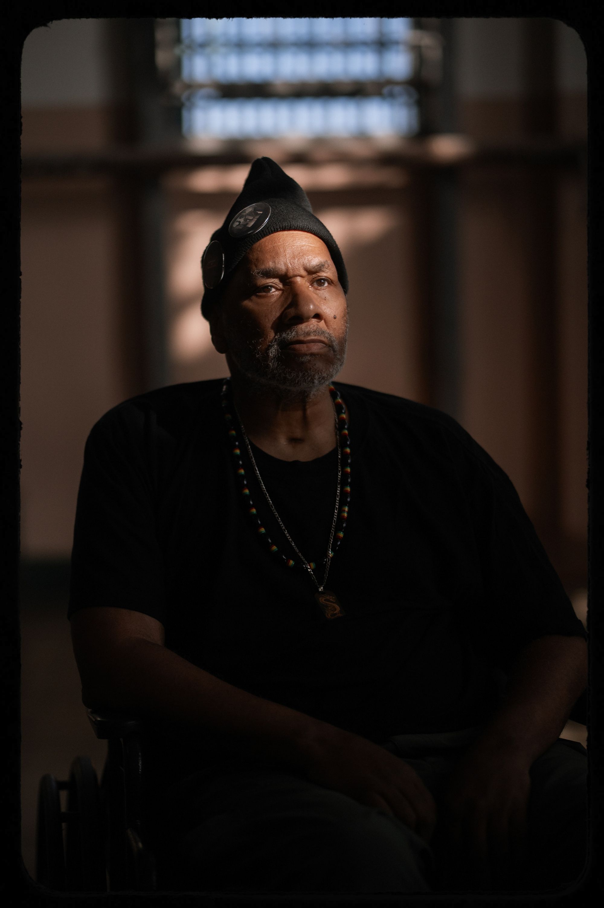 A photo shows Sitawa Jamaa, a Black man wearing a black beanie and a black T-shirt, sitting in a wheelchair in an indoor space. 