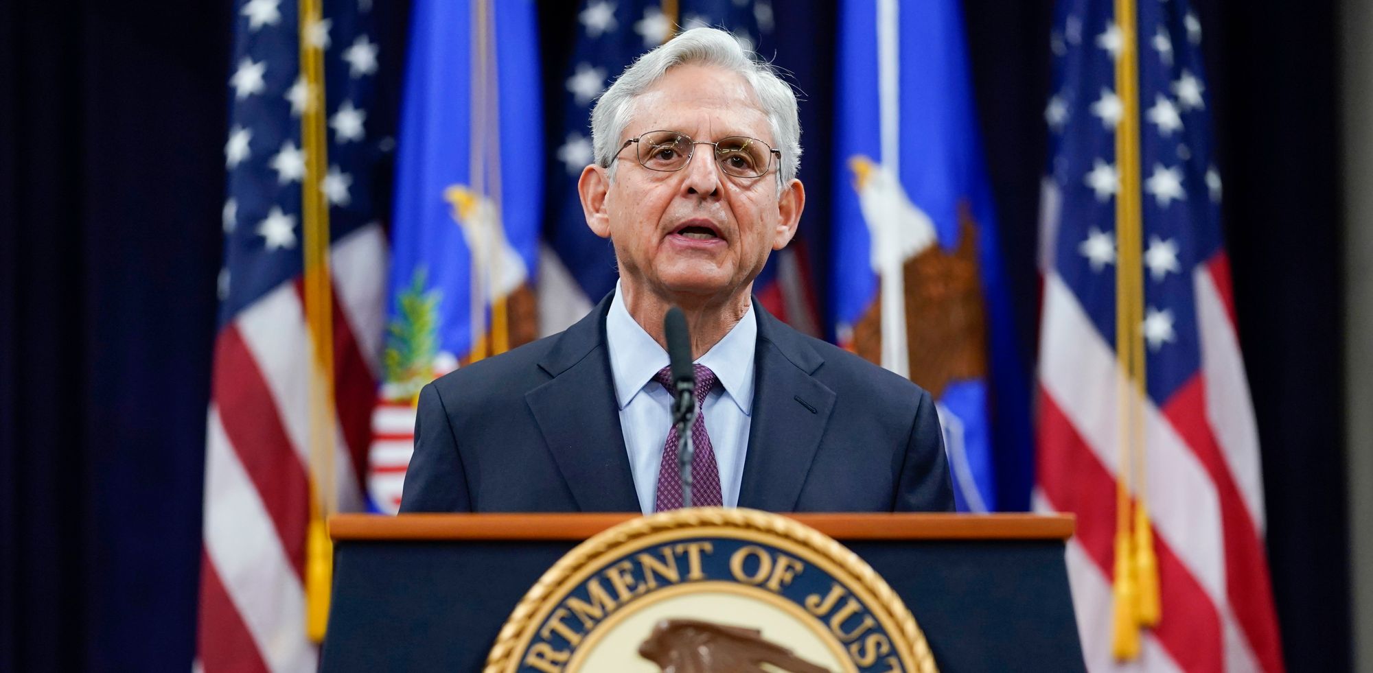 Merrick Garland, a man with light-toned skin and white, short hair, stands at a podium with a backdrop of American flags. 