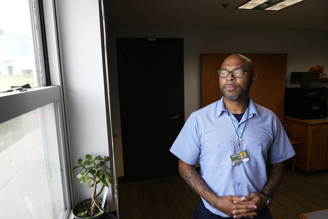 A photo shows Steve Grant, a Black man wearing a pale blue T-shirt that is part of a uniform, standing near a window. He has a badge hanging around his neck. 