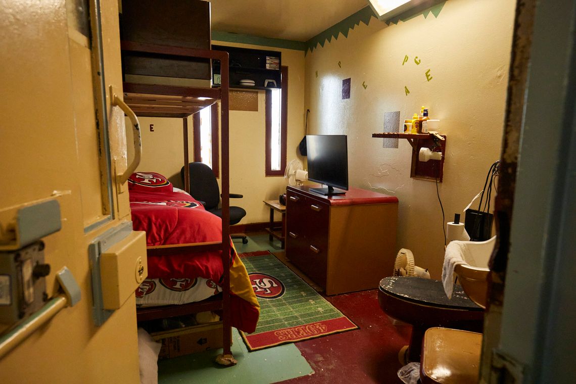 A small room with two narrow, vertical windows has a cabinet with a flat-screen television sitting on top of it on the right side. On the left is a bed with a red blanket with the San Francisco 49ers logo on it. A toilet and small sink sit in the near right corner.