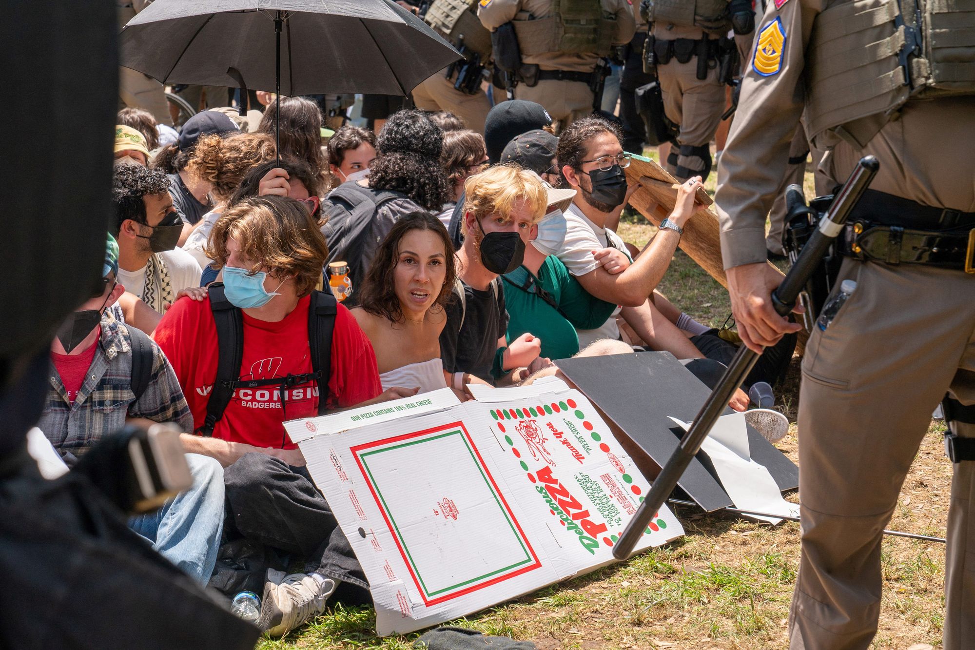 A photo shows a group of people sitting in a circle. In the foreground, a Texas State trooper holds a baton. 