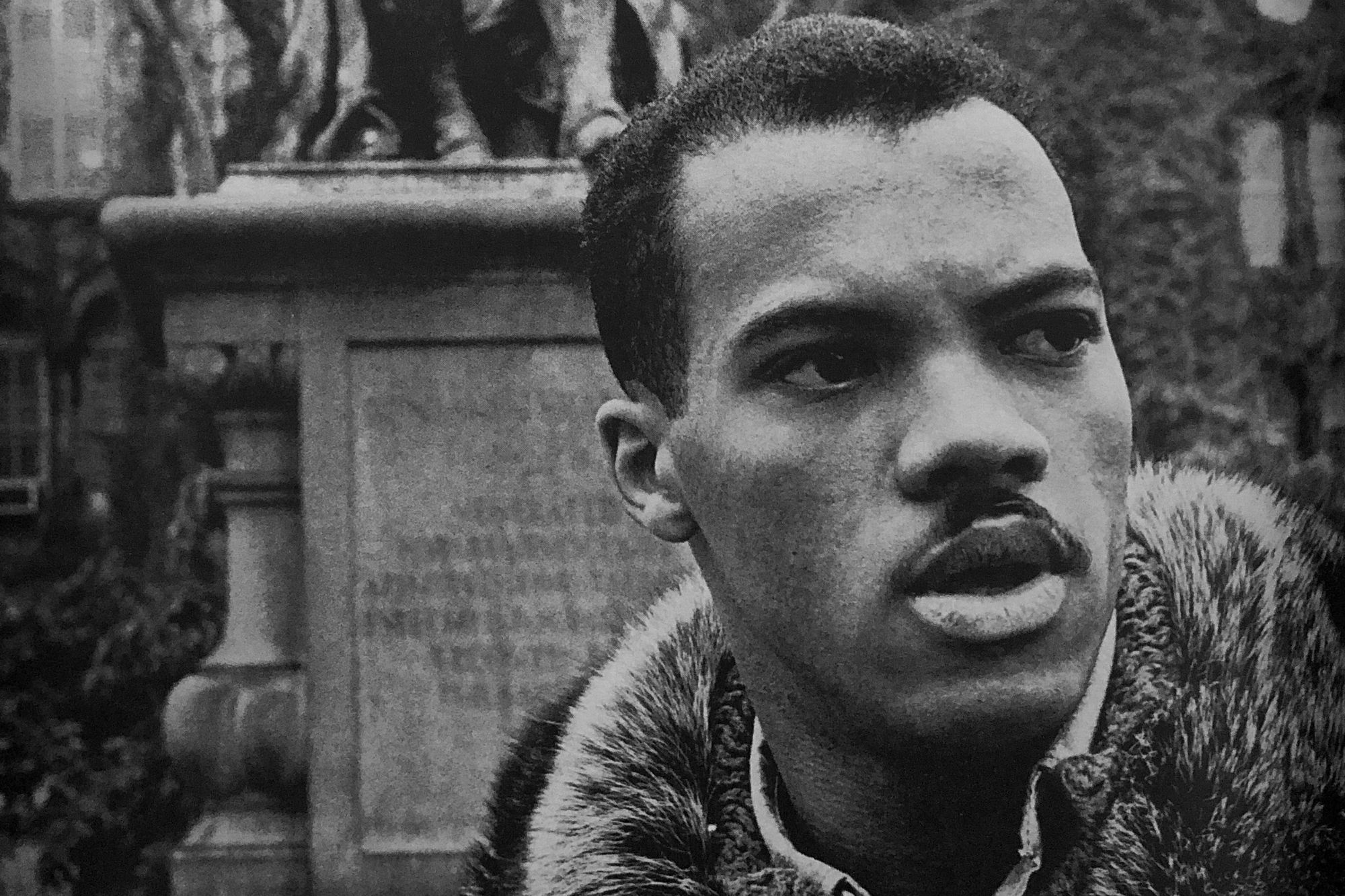 A black and white image of a Black man in a button down shirt and fur-collared jacket, standing in front of a statue.