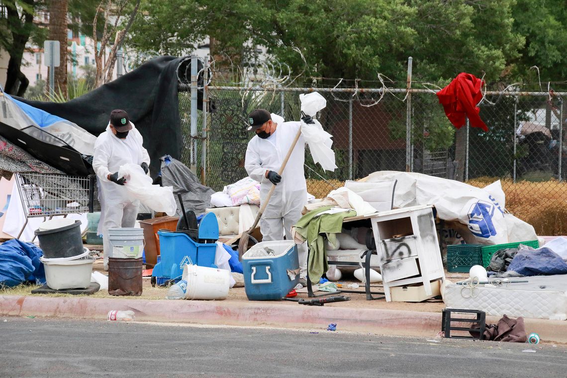 Two city workers, wearing black caps, black gloves and white coveralls, hold plastic bags as they “sweep” the remains of a homeless encampment.