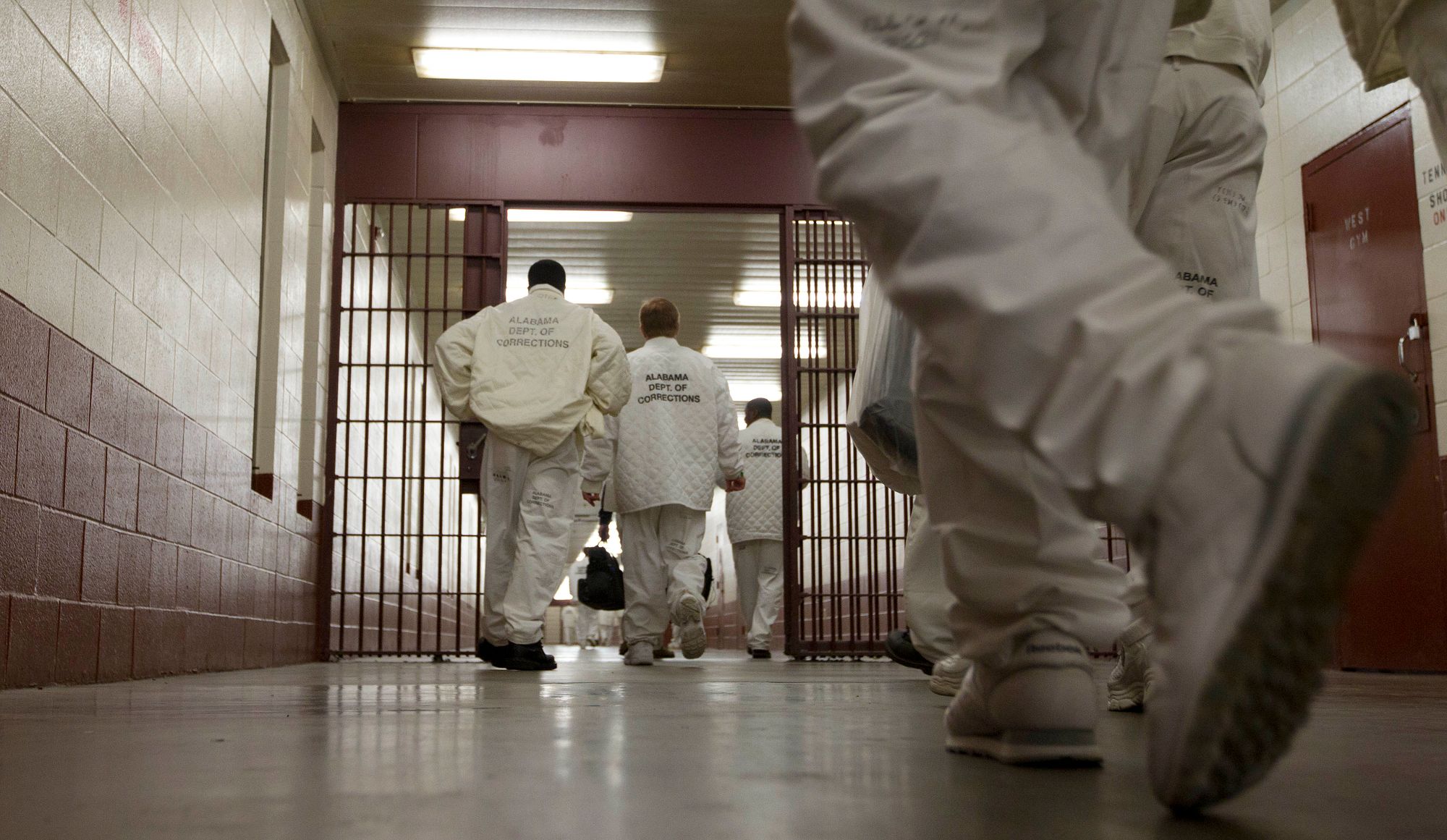 Prisoners, with their backs facing the camera, walk past a gate in a hallway. The men are dressed in white uniforms with the words "Alabama Dept. of Corrections" on their backs.

