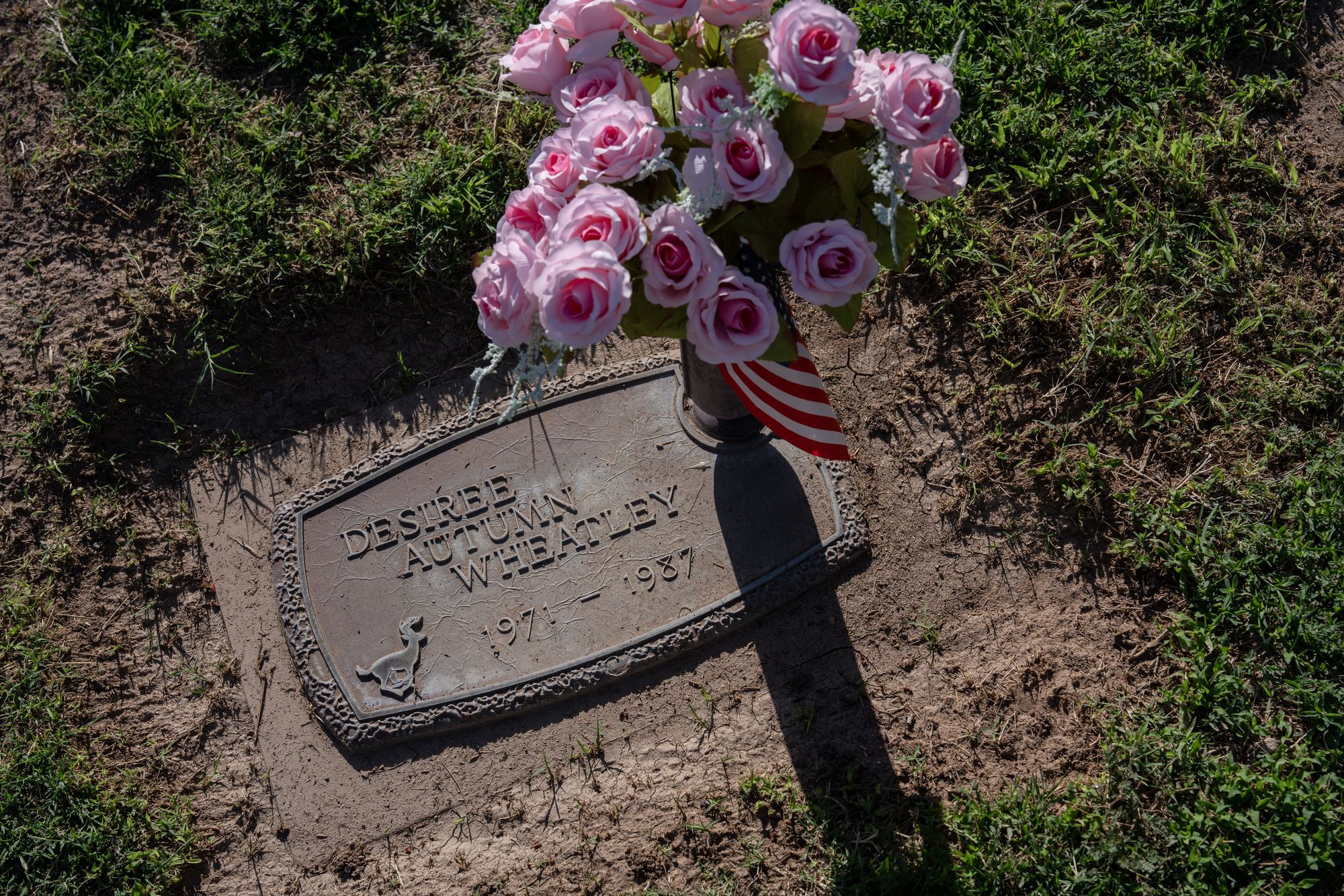A photo shows a grave plaque on the ground that reads, “Desiree Autumn Wheatley 1971-1987.” Placed on top of the grave are a small American flag and a bouquet of pink flowers. Grass surrounds the grave. 