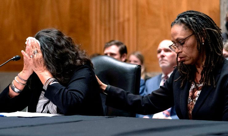A White woman covers her face with both hands as she cries during a court hearing. A Black woman on the right consoles her with her hand on her back.