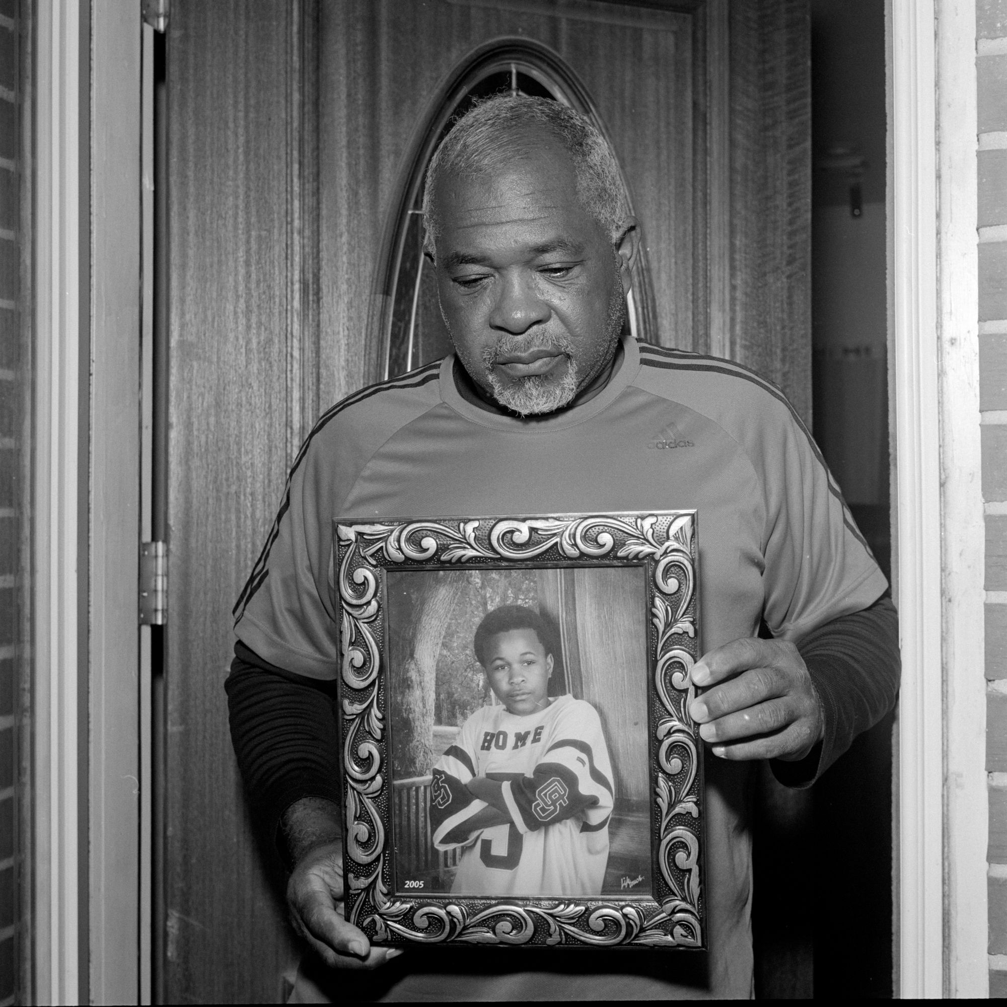 A Black man wearing a T-shirt over a long-sleeved shirt holds a framed picture of his son when he was a small boy. His son is wearing a football jersey in the photo. 