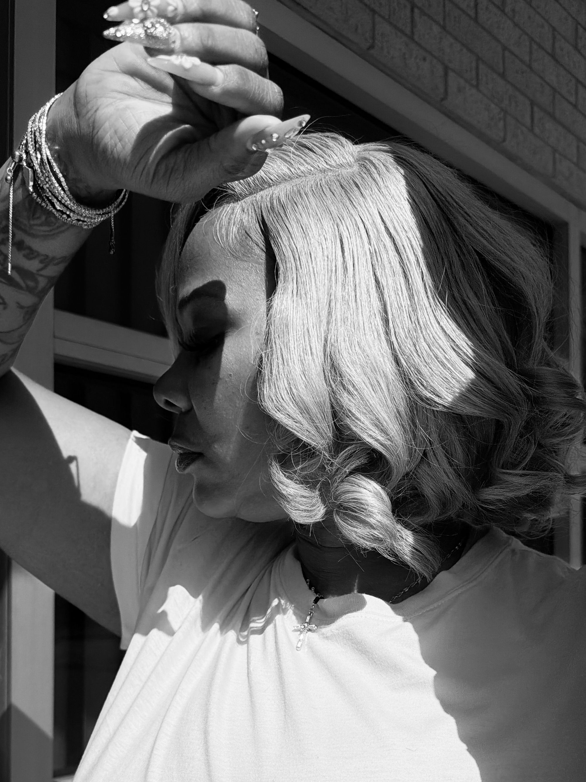 A black-and-white photo shows a Black woman with curled, blonde medium-length blonde hair in profile, looking to the left. She has the back of her hand resting on her head. 