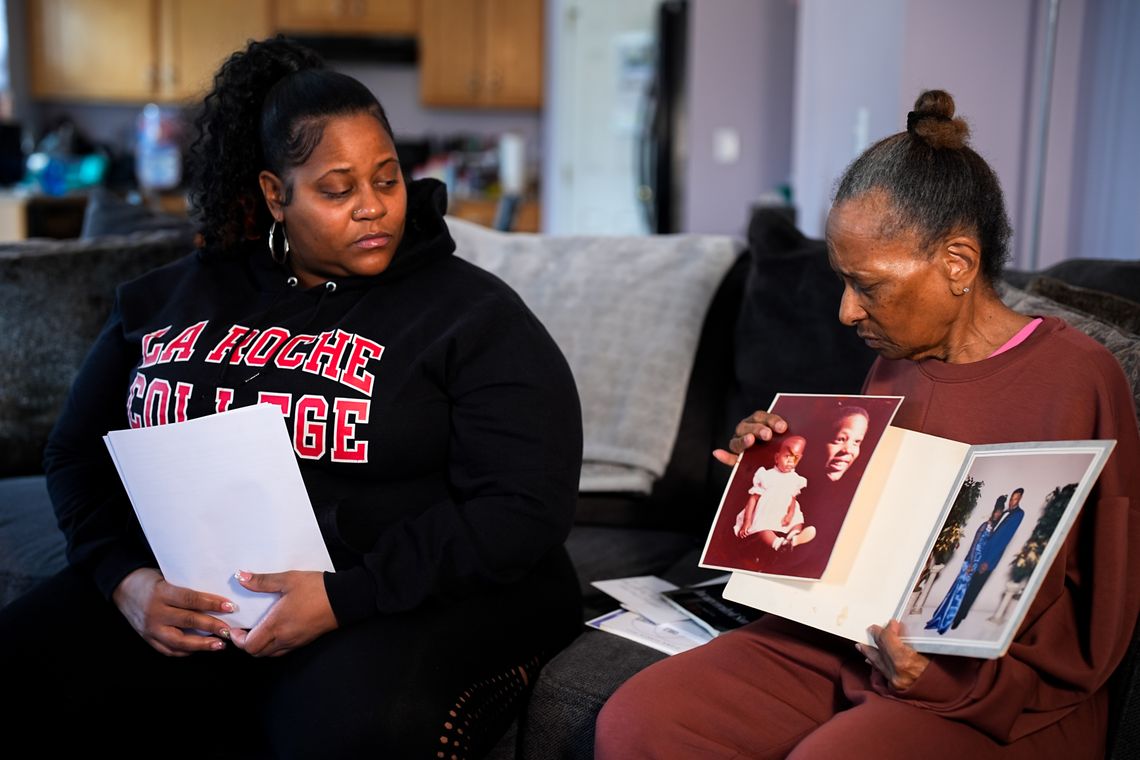 A photo shows Amber Hall, left, a Black woman wearing a black sweatshirt that reads “La Roche College” holding some white sheets of paper. Her mother, Cora Fleeta Hill, right, a Black woman wearing a maroon matching sweatshirt and sweatpants, holds some photos showing her son Jayson Murphy while they sit on a couch. 