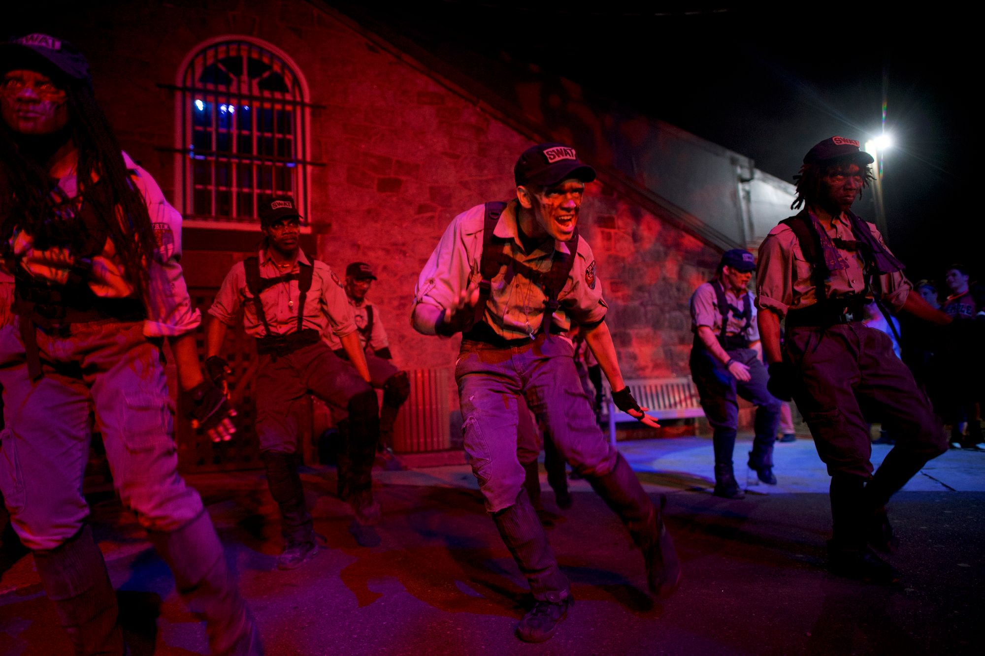 Six actors are dressed in matching prison uniforms and with zombie make-up. A magenta light shines on them. 