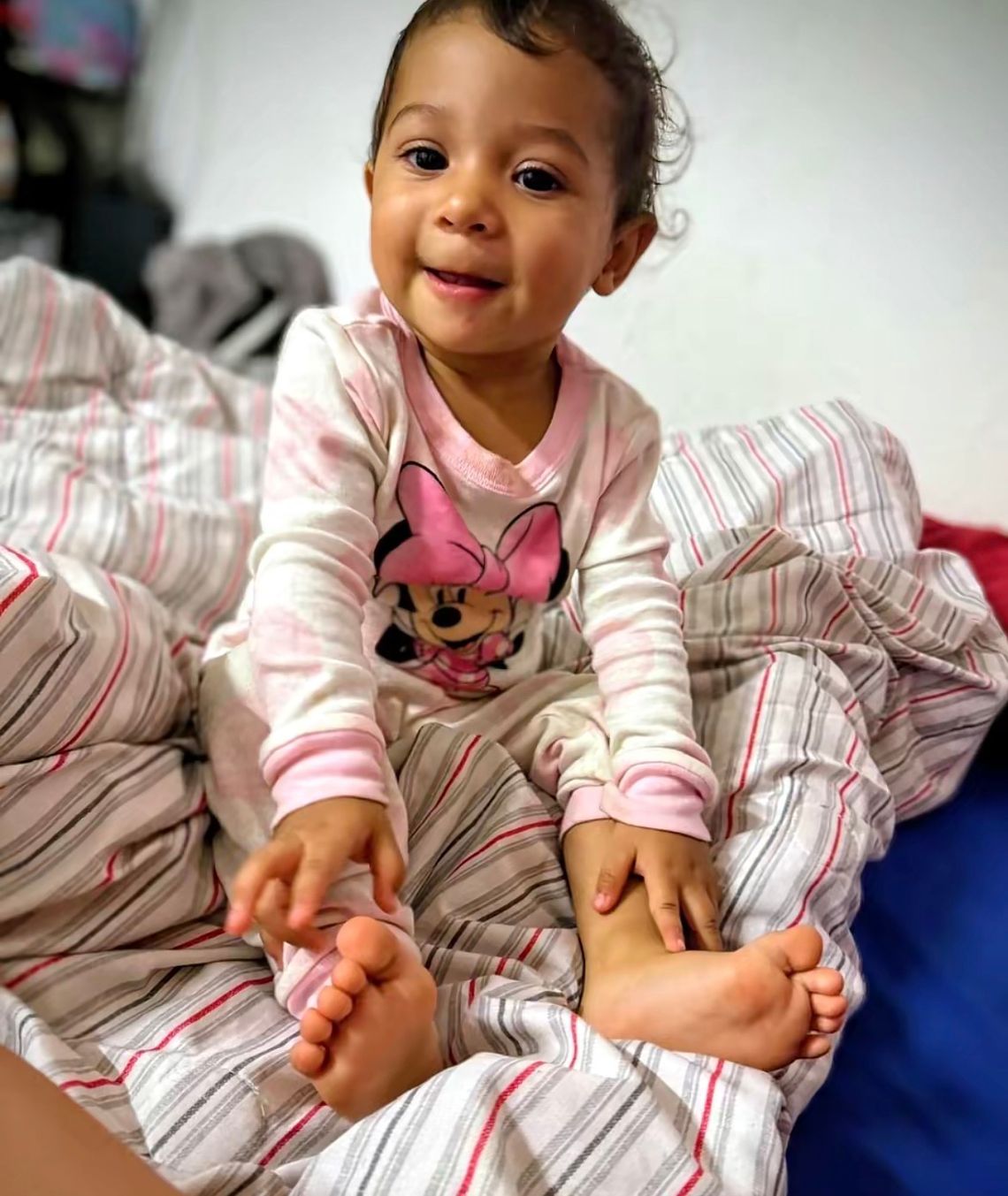 A photo shows a baby with brown hair and medium-toned skin, wearing a Minnie Mouse pajama set, sitting on a bed. 
