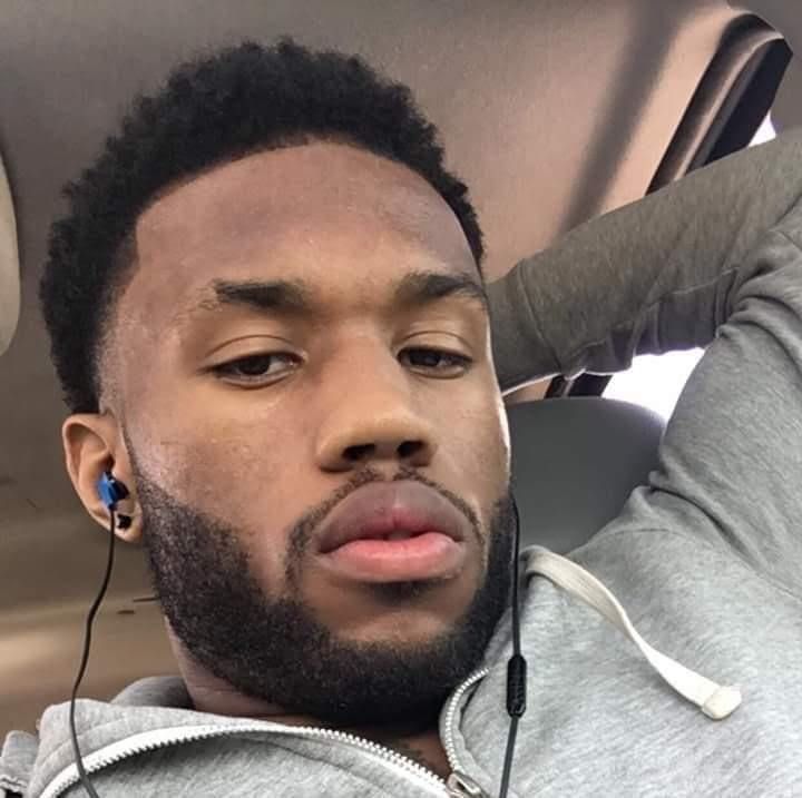 A black man with short hair and a gray hoodie looks at the camera.
