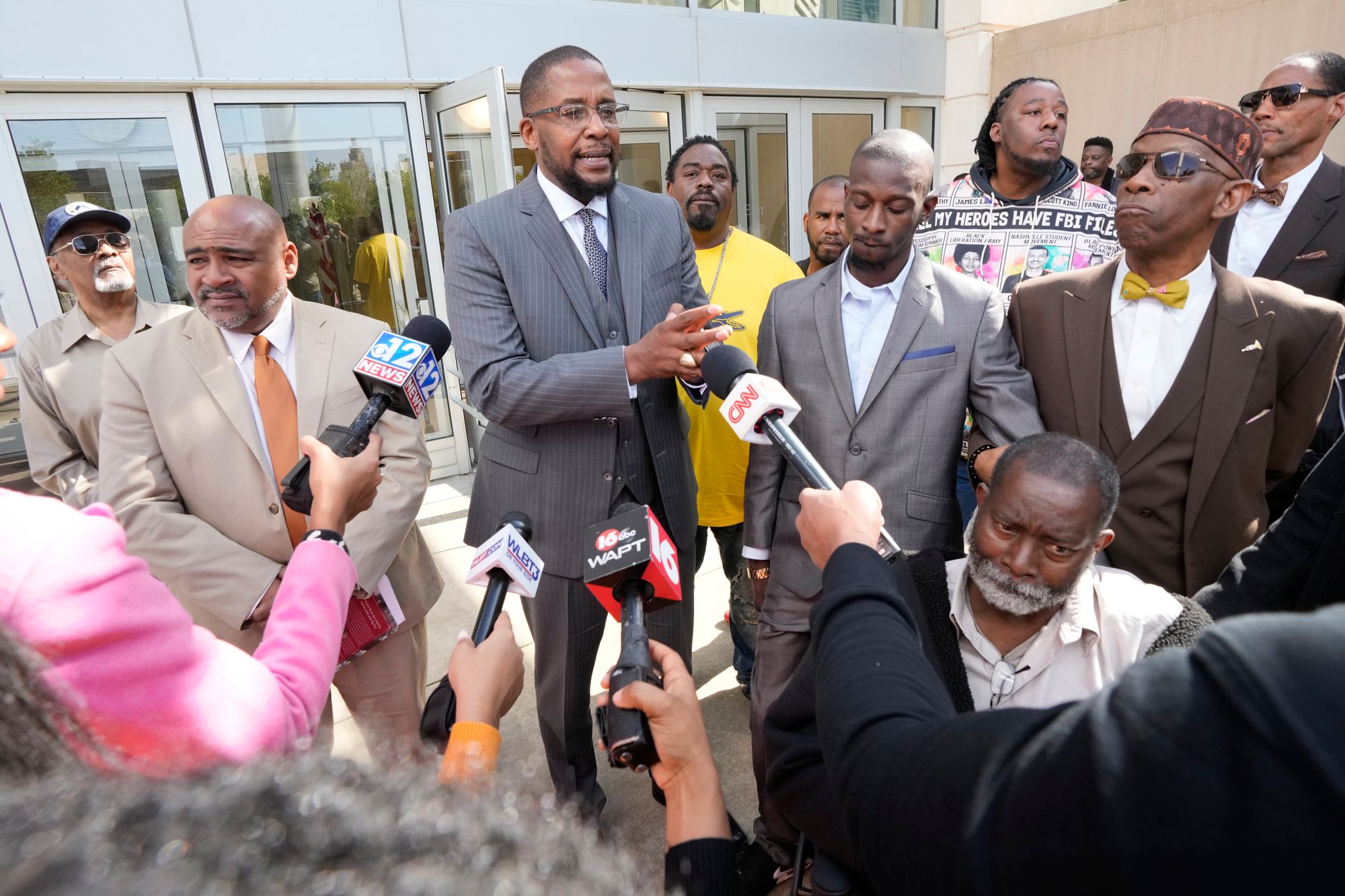 Malik Shabazz, a Black man in a gray suit, speaks to reporters, as Michael Corey Jenkins, a Black man in a gray suit, stands next to him. A group of other people stand near them, in front of microphones held up by reporters. 