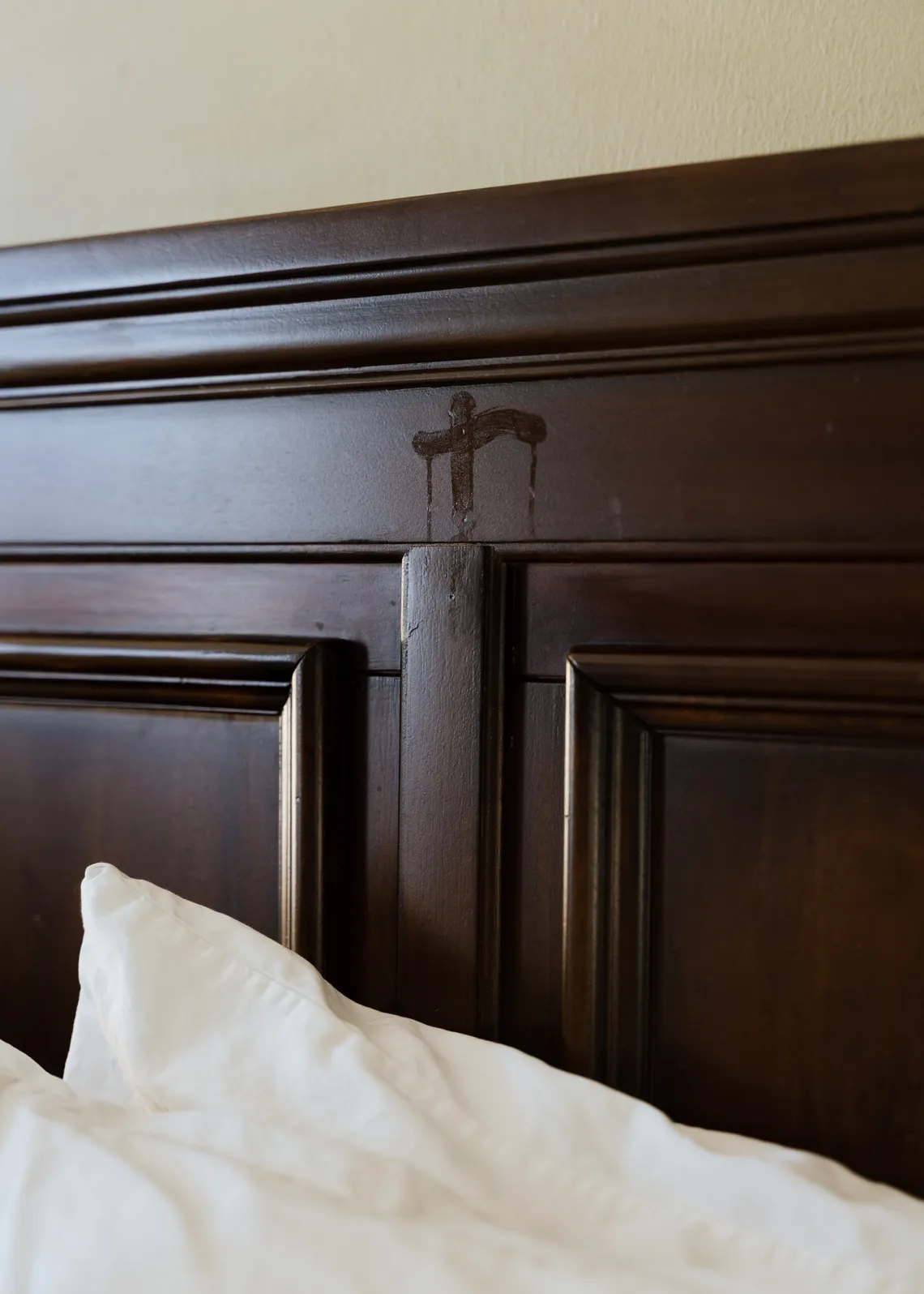A photo shows a brown wooden headboard of a bed marked with olive oil in the shape of a cross. A white pillow is at the bottom of the photo. 