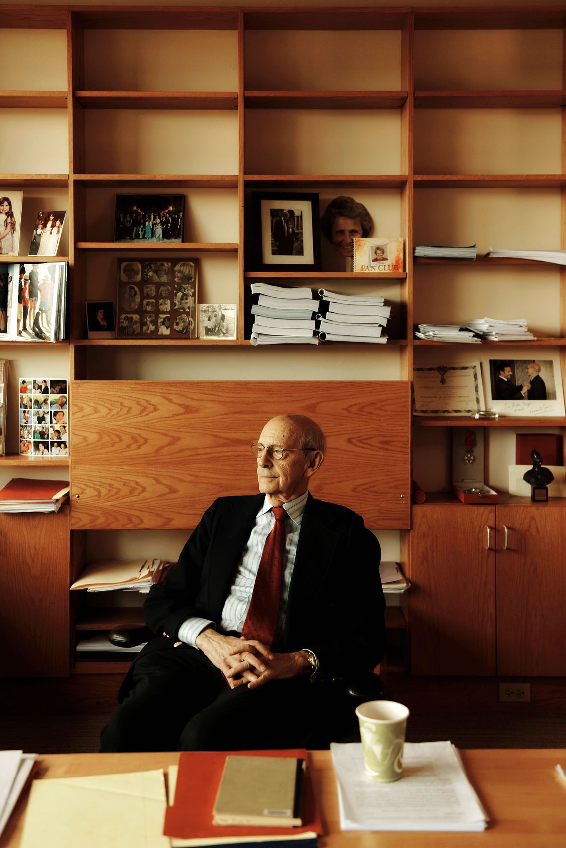 An older White man wearing glasses, a black suit, striped shirt and red tie, sitting in front of his office desk. 