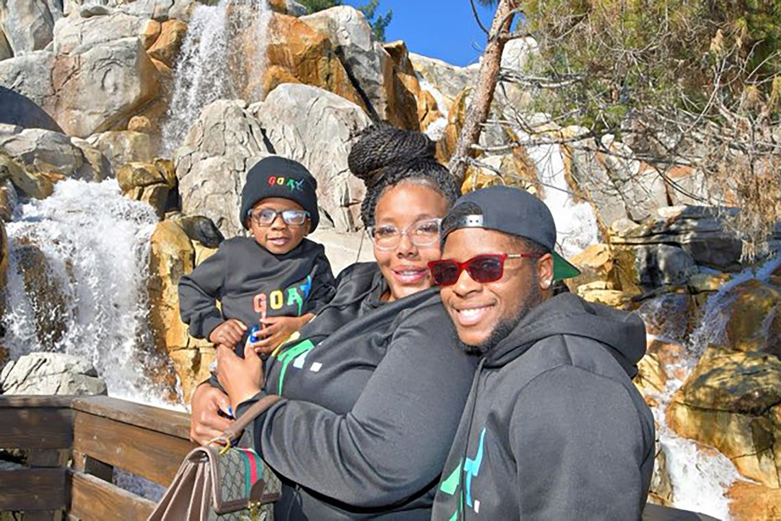 A Black woman with glasses and a braided bun smiles for a photo while holding a Black baby wearing glasses and a beanie. The woman is standing next to a Black man in sunglasses and a backwards baseball cap. 

