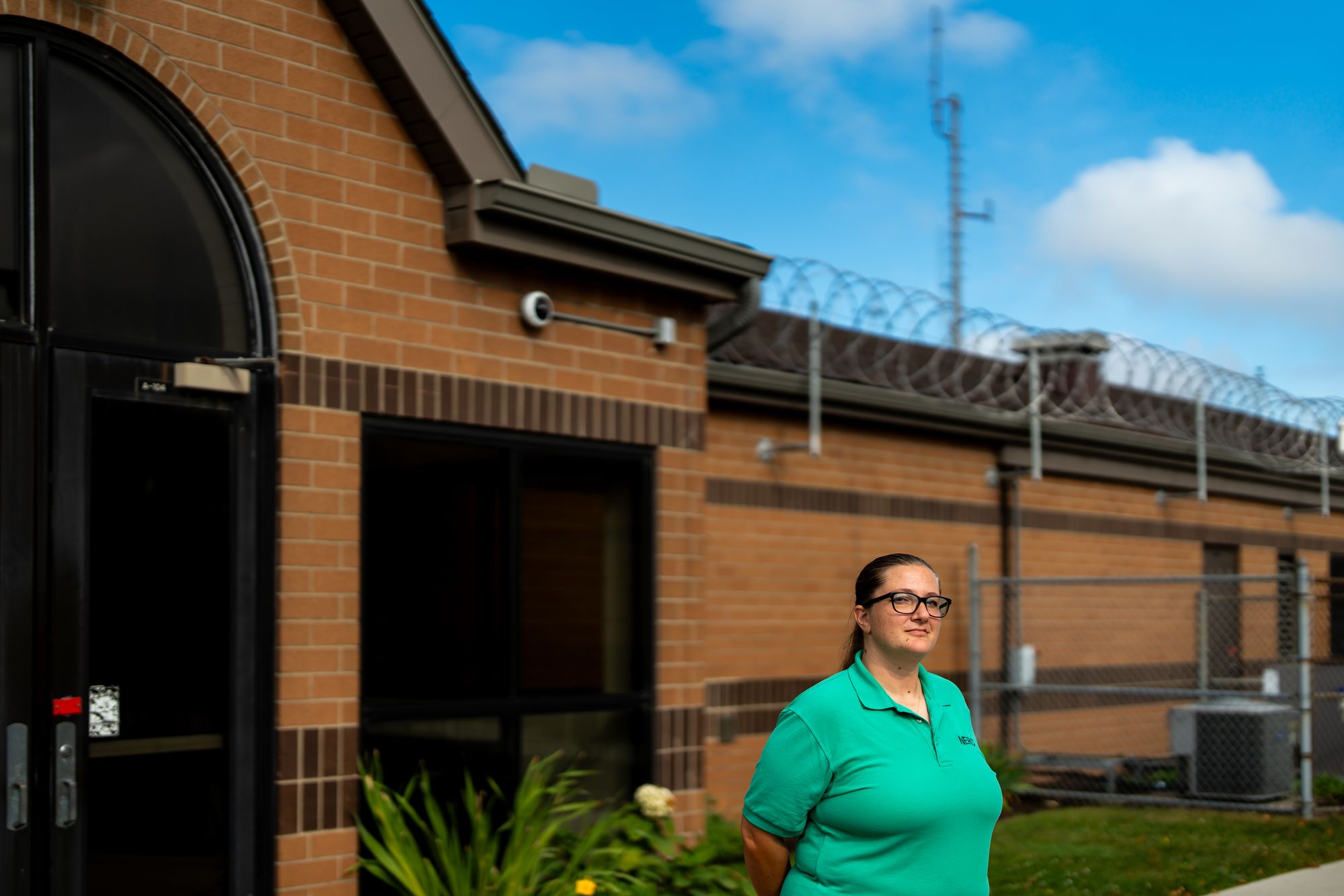 A photo shows Barbara Devine, a White woman with glasses wearing a green T-shirt that is part of a uniform, standing outdoors in front of a brown brick building.  Barbed wire fencing is visible above the building to the right.  