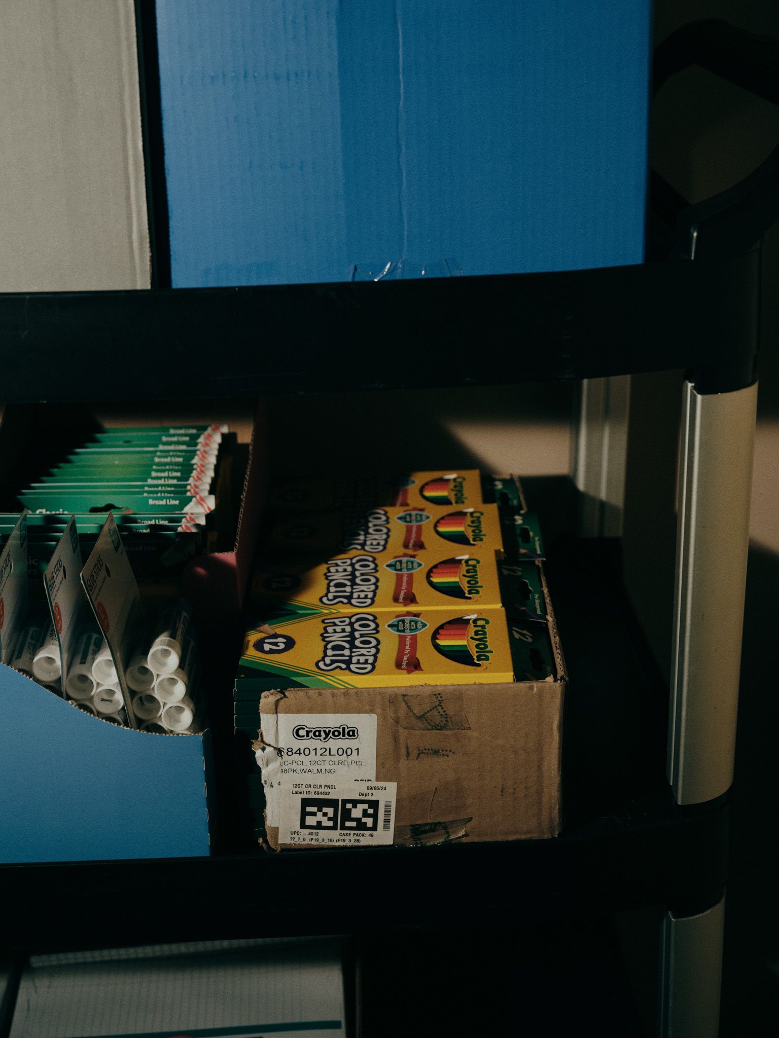 Shelves hold a box full of packages of colored pencils, a box of glue sticks, and a box full of packages of markers.
