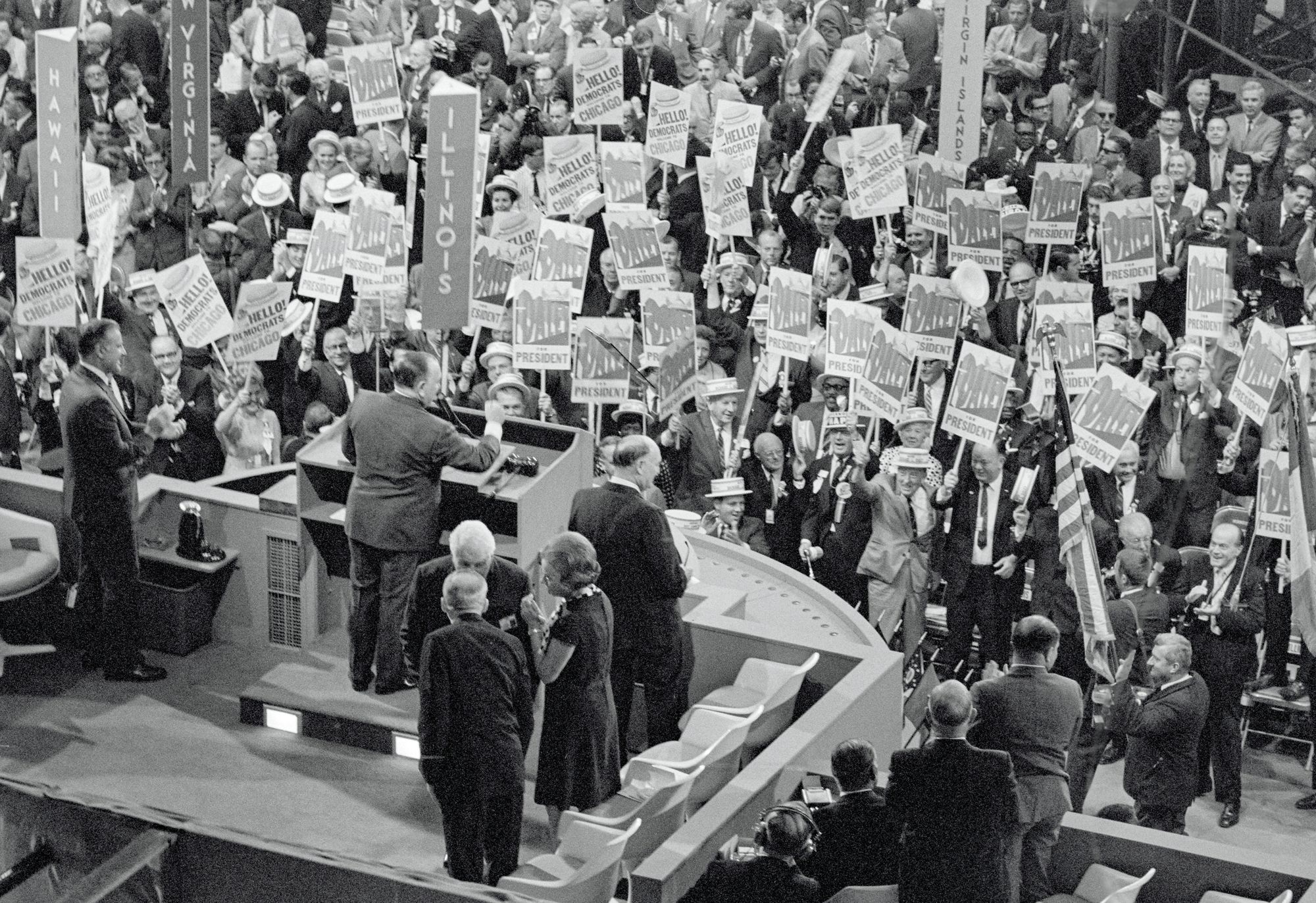 A black and white photo shows a White man in a suit on a raised platform delivering a speech to a crowd.