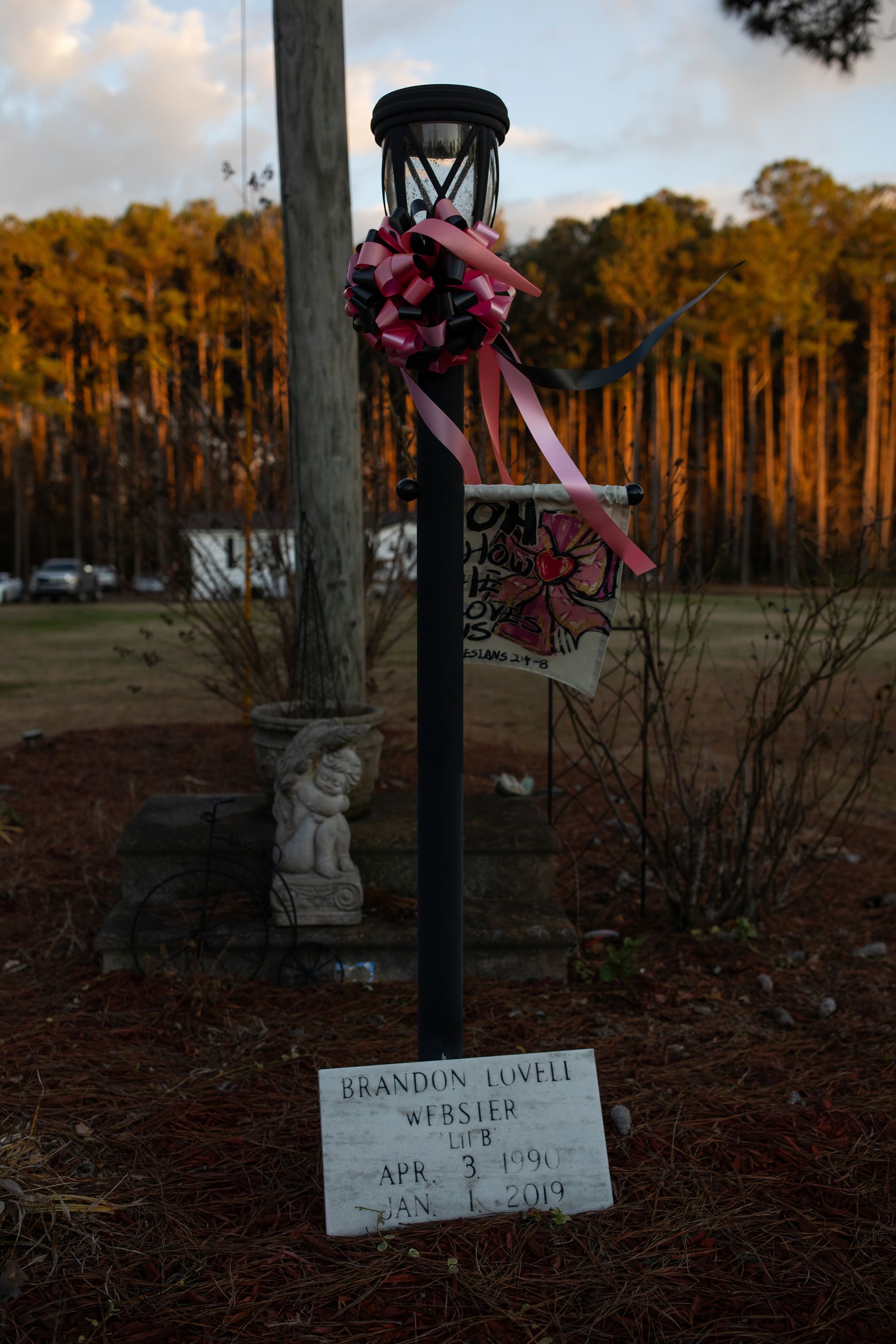 A photo shows a memorial that is marked by a stone that has “Brandon Lovell Webster, April 3, 1990-Jan. 1, 2019” carved into it. A lightpost also marks the memorial and has pink and black ribbons tied on it. 