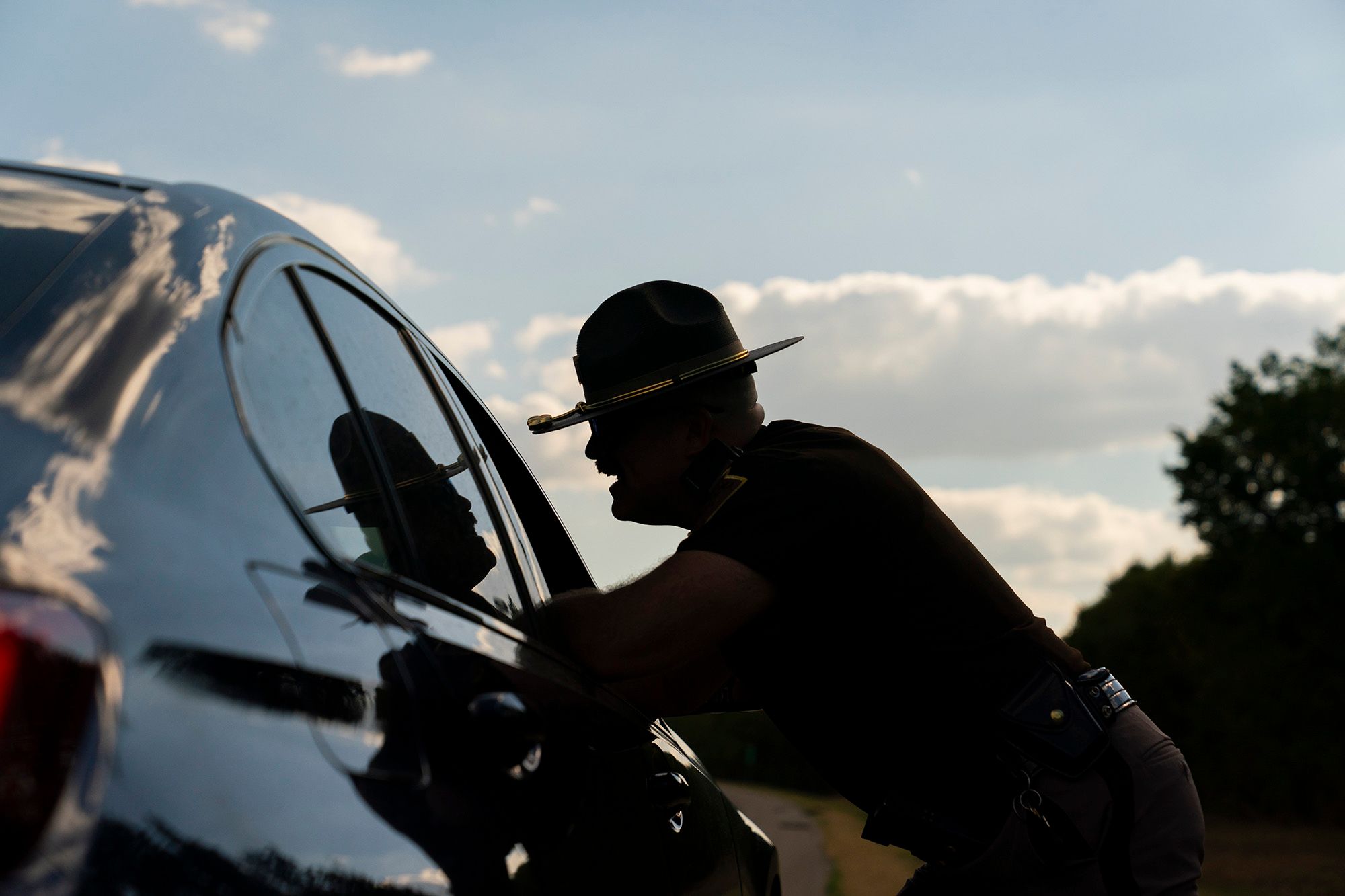 A photo shows a silhouette of an trooper talking with a driver who was pulled over. The trooper is leaning on the car with an open window. 