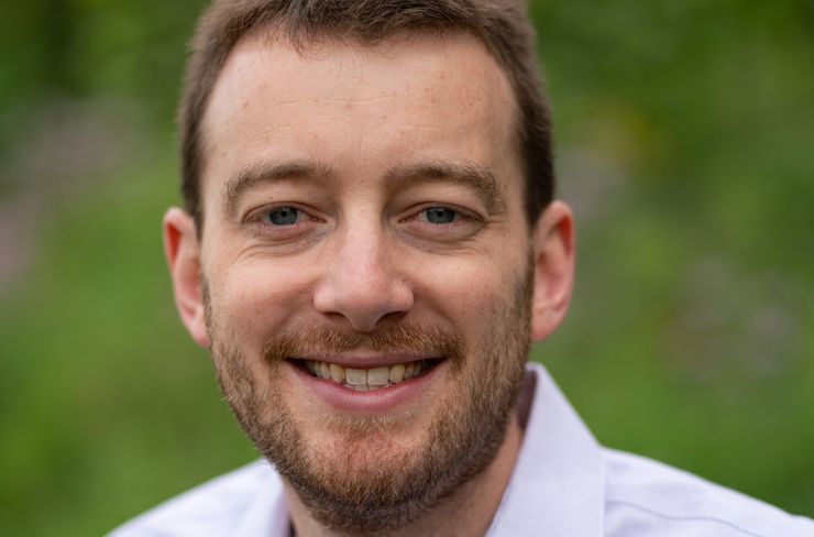 A photo shows a headshot of a man with smiling White man and a beard standing outdoors.  