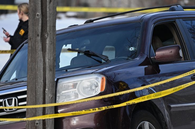 A bullet hole appears in the bottom left of the windshield of a dark-colored SUV that has struck a pole. In the foreground are two ribbons of police tape and in the background stands a person with a phone. 