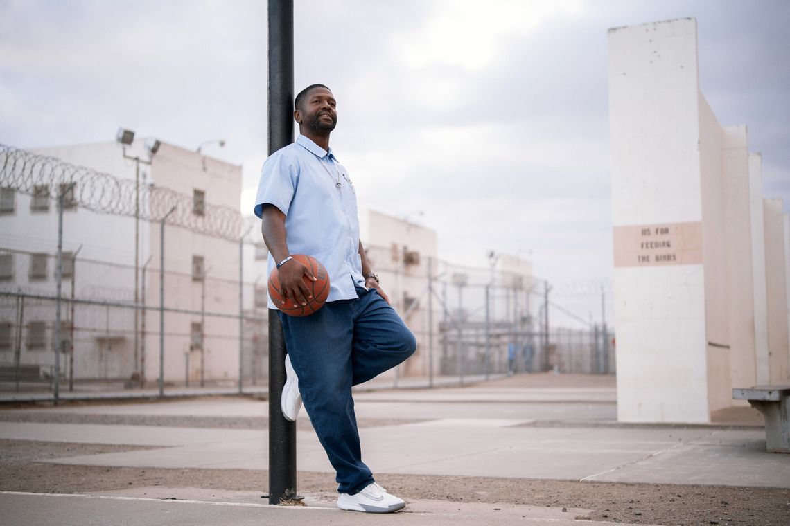 A photo shows a man with dark-toned skin and wearing a light blue shirt and dark blue pants, leaning against a pole while holding a basketball on the prison yard. 